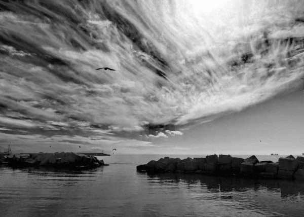 Suggestiva fotografia in bianco e nero scattata dal lungomare del centro storico di Salerno. Si vedono gli scogli che proteggono il lungomare dalle onde del mare agitato e il cielo con una formazione di nubi cirriformi che creano un'atmosfera particolare e sembrano di allargare l'orizzonte.