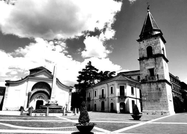 Fotografia in bianco e nero con un'atmosfera drammatica. La piazza nella foto si trova a Benevento in Campania. Il grandangolo permette non solo di inquadrare tutta la piazza con la chiesa, il campanile e l'obelisco ma crea una prospettiva particolare in cui tutti i monumenti sembrano di allungarsi verso il cielo. Fotografia di Annett Siviglia-Mann.