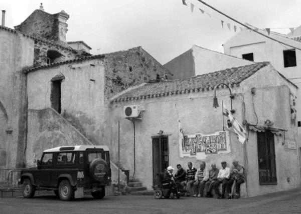 Questa fotografia del genere Street Photography mostra un gruppo di anziani seduti davanti ad una casa in una piazza nel centro storico di Cagliari in Sardegna. Una scena tipica per l'Italia dove i pensionati maschi si incontrano nella piazza del paese, si siedono insieme e fanno due chiacchiere mentre le mogli fanno i servizi a casa.