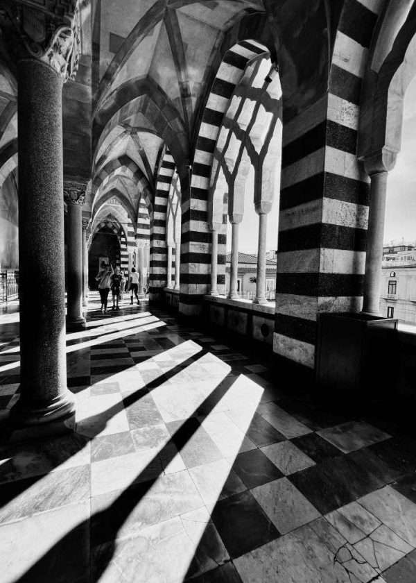 Una foto insolita del Duomo di Amalfi che è famoso per la lunga scalinata che qua non si vede. Questo scatto è stato fatto nell'atrio prima di accedere all'interno del duomo. Questa vista è riservata a chi si prende la briga di salire tutte quelle scale prima. Una fotografia in bianco e nero che gioca con la luce del sole che entra dai portici e forma ombre contrastanti che si ritrovano con il pavimento e le mure decorate in bianco e nero sempre.
