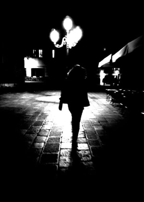 In questa fotografia in bianco e nero si vede la sagoma di una donna che cammina per le strade di Venezia in piena notte. La piazza è illuminata da un lampione che dona poca luce. Street Photography di Annett Siviglia-Mann.