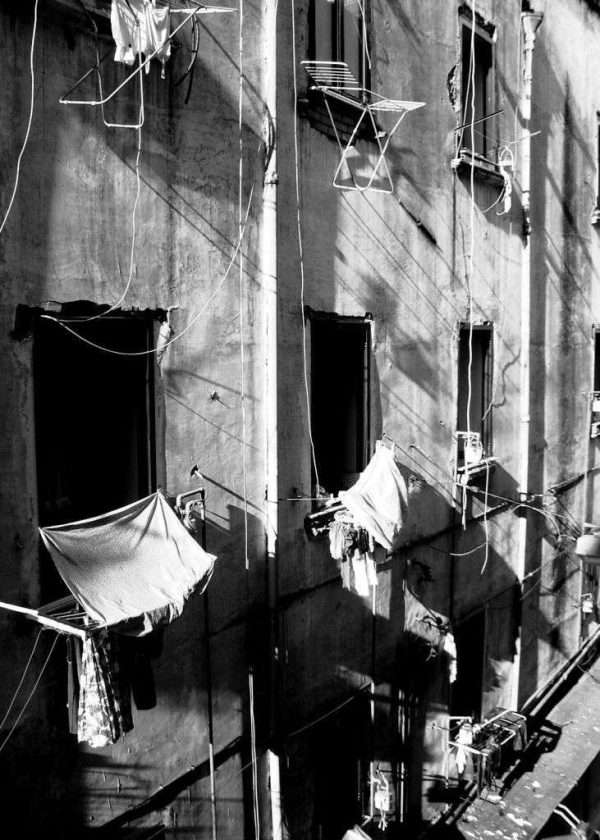 Una foto di una facciata nel centro storico di Napoli. La fotografia in bianco e nero mostra un palazzo un po' degradato con gli stendini per la biancheria attaccati fuori alle finestre che non hanno nemmeno un telaio decente e neanche i fili elettrici sono a norma. Ma non importa, perché se vivi a Napoli sei felice a prescindere.